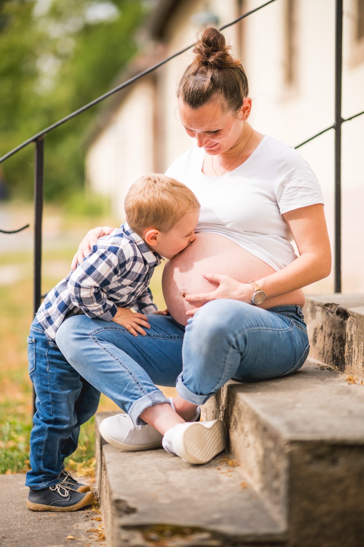 Babybauch-Shooting – Geschwisterkind küsst den Bauch der schwangeren Mutter auf einer Treppe