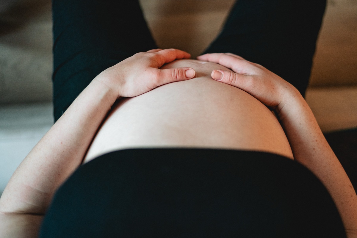 Babybauch-Shooting zuhause – schwangere Frau in dunkler Kleidung, Hände ruhen sanft auf dem Bauch, stimmungsvolles Licht