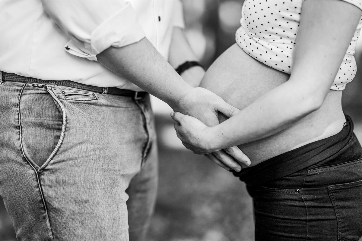 Babybauch-Shooting – Detailaufnahme: Hände eines Paares zärtlich verschränkt am schwangeren Bauch, Schwarzweißfoto