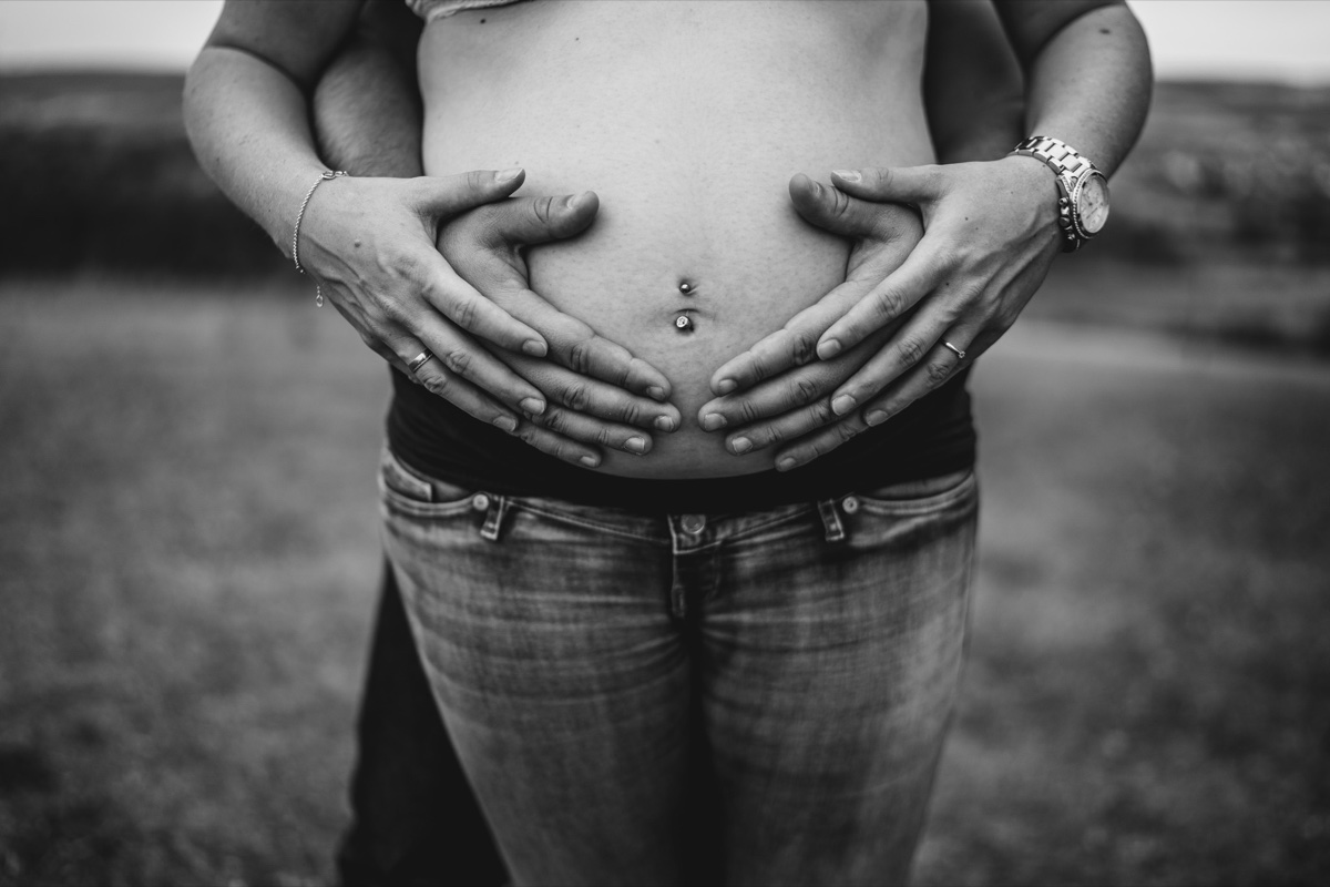 Babybauch-Shooting – vier Hände eines Paares halten liebevoll den schwangeren Bauch, Schwarzweißfoto