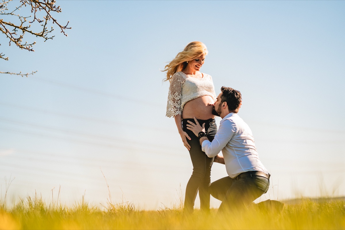 Babybauch-Shooting – Mann kniet und küsst liebevoll den Bauch, Paar auf einer Wiese im warmen Abendlicht
