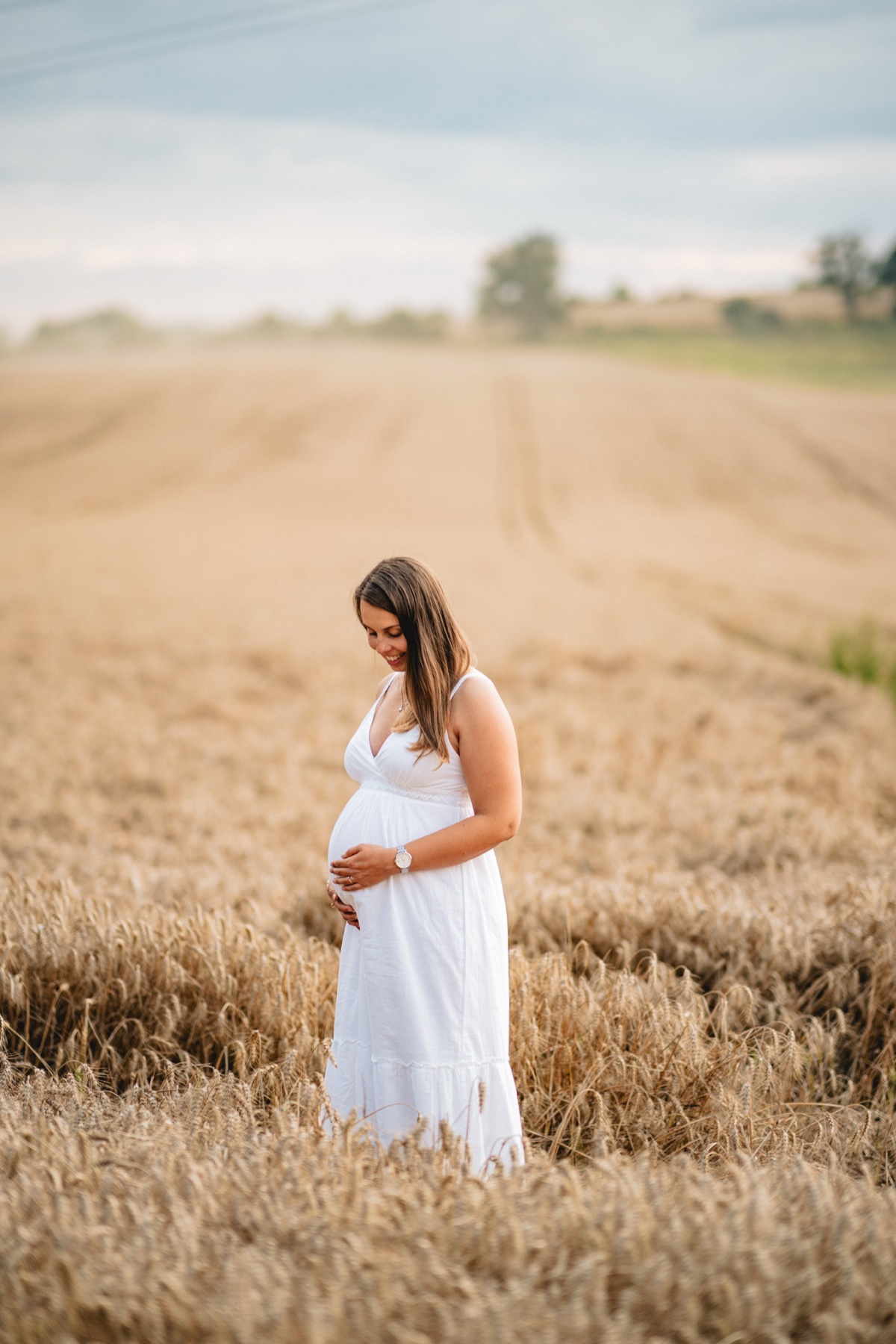 Babybauch-Fotoshooting – schwangere Frau im weißen Kleid blickt lächelnd auf ihren Bauch, goldenes Weizenfeld im Hintergrund