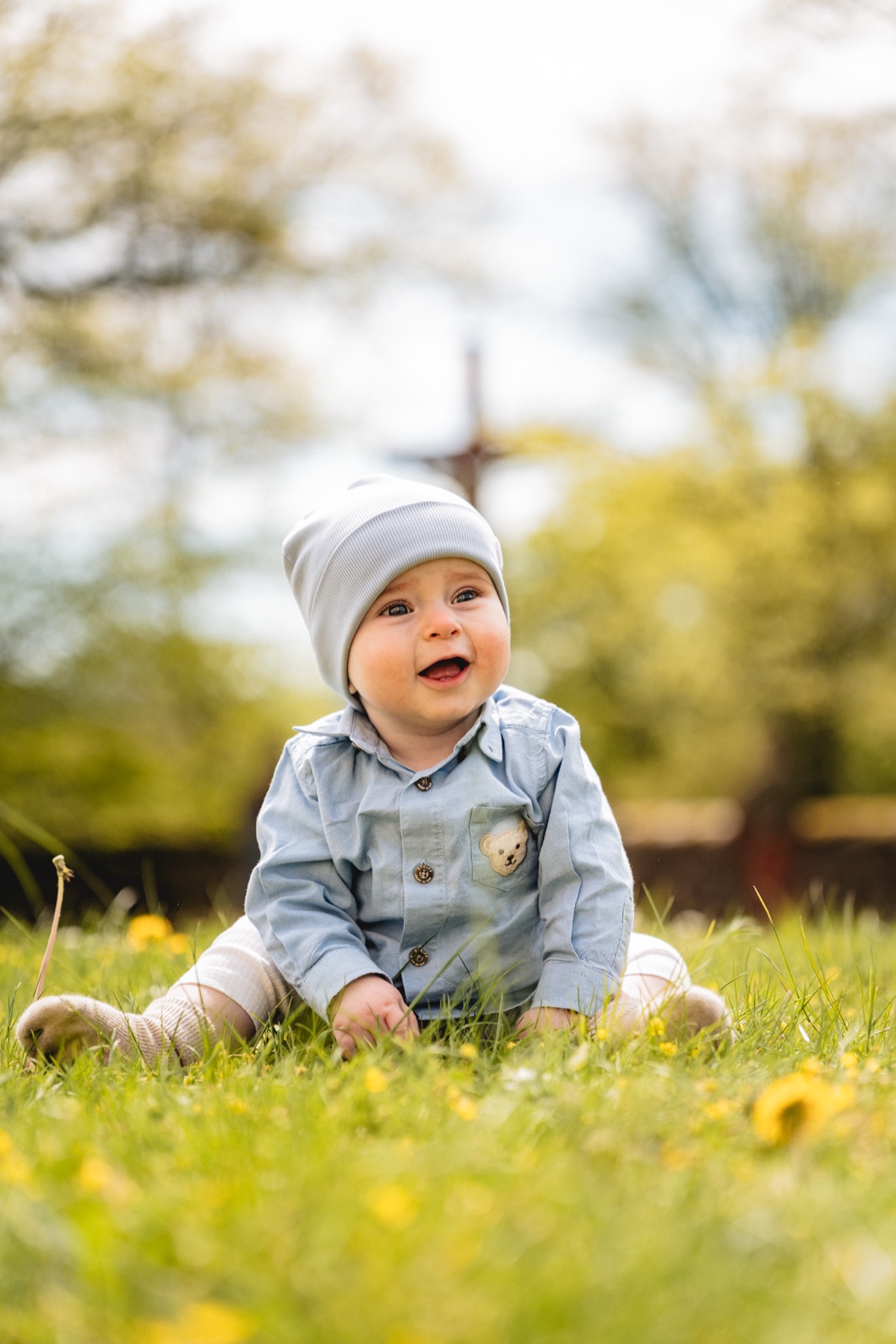 Baby-Portrait – lachendes Baby mit Mütze sitzt auf einer Blumenwiese im Frühling