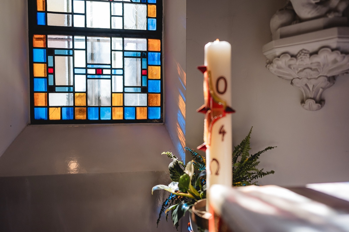Taufe Fotografie – buntes Kirchenfenster mit Osterkerze und Grünschmuck, Lichtspiel in der Kirche