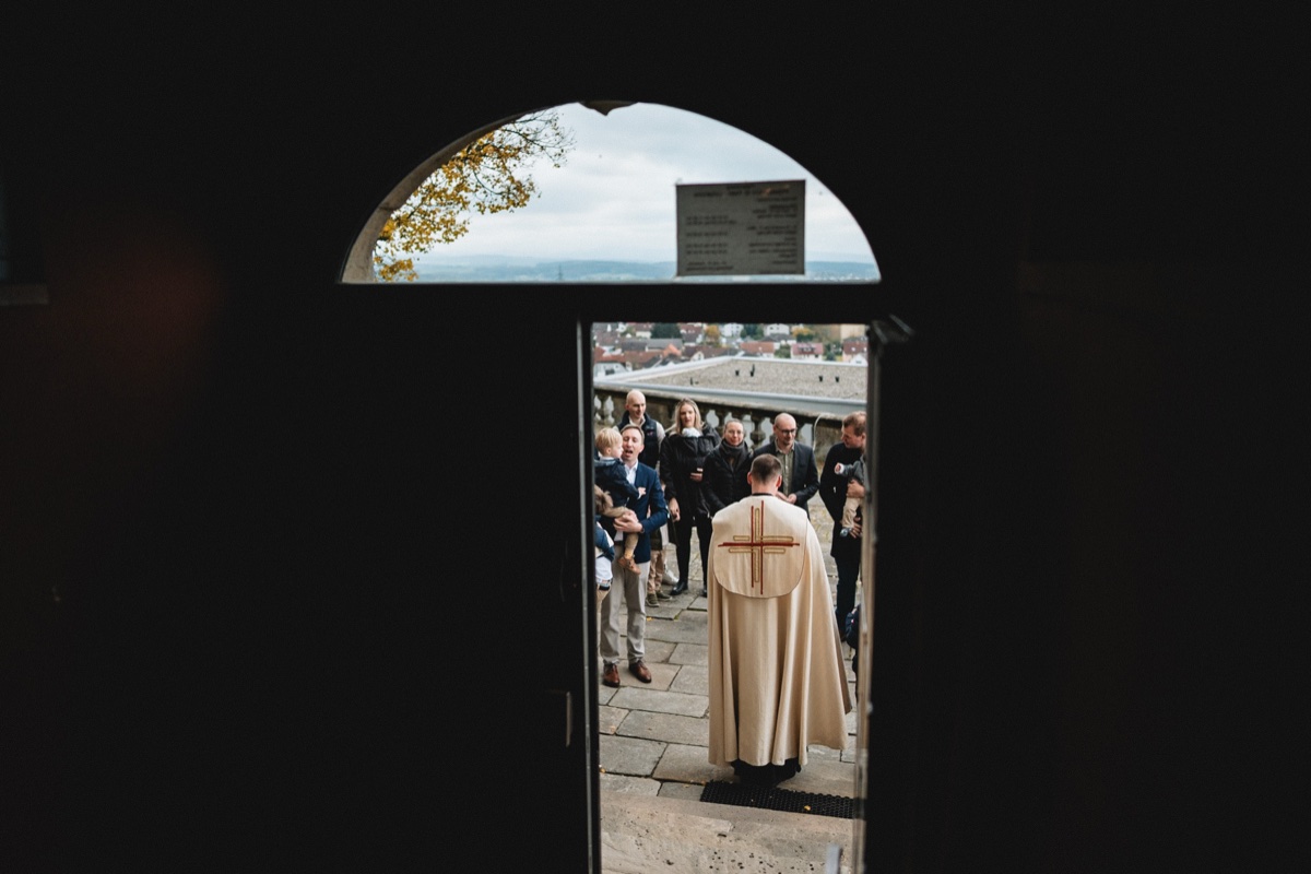 Taufe Fotograf – Blick durch den Kirchenbogen auf Familie und Priester beim festlichen Einzug zur Tauffeier