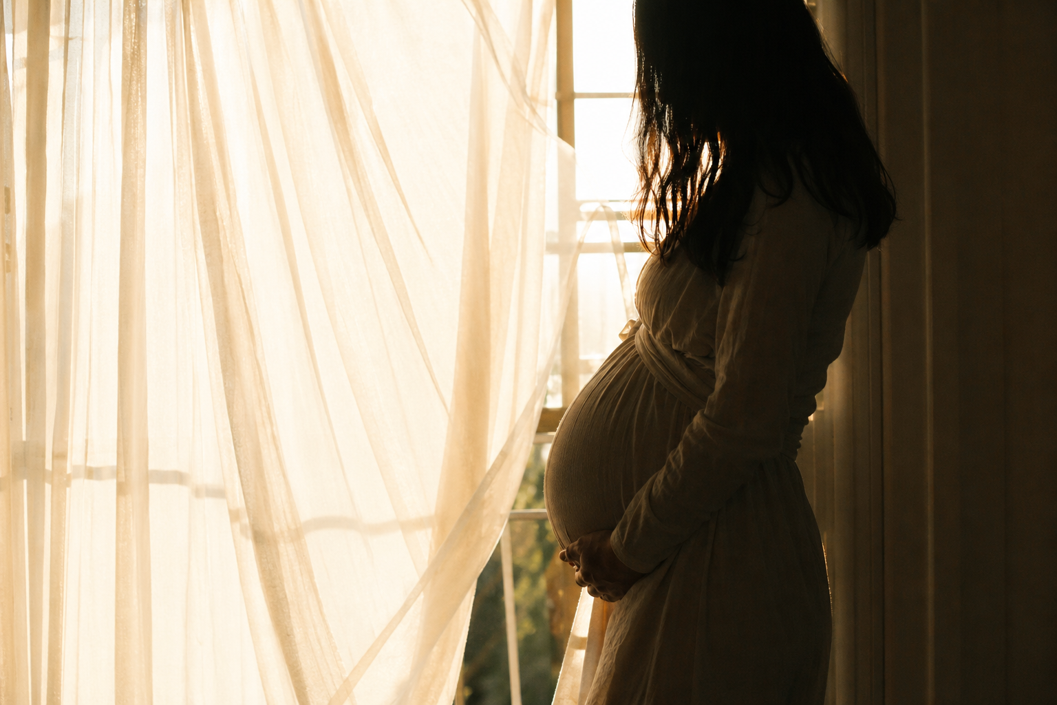 Schwangere als Silhouette vor einem hellen Fenster, eine Hand auf dem Bauch, weicher heller Vorhang seitlich, warmes Gegenlicht